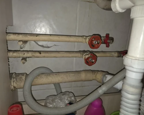 Instalație sanitară veche aflată sub o chiuvetă, formată din țevi metalice vopsite cu robineți clasici uzați cu rozetă roșie, alături de o țeavă de scurgere din PVC și furtunuri flexibile de evacuare.