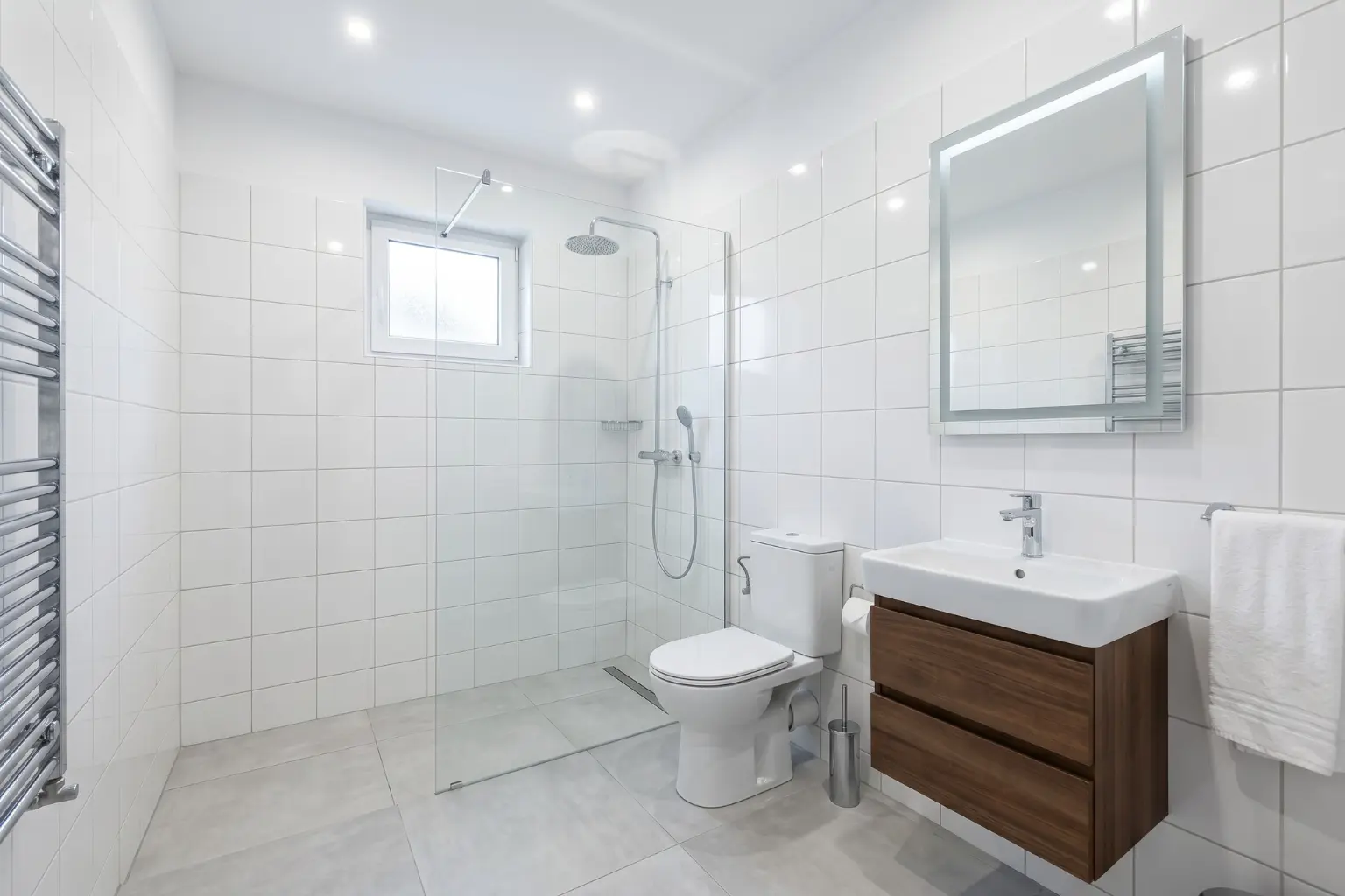 O baie modernă, recent renovată, cu faianță albă, dotată cu o cabină de duș de tip walk-in cu paravan din sticlă, vas WC, lavoar cu mobilier suspendat din lemn, oglindă iluminată cu LED și un calorifer portprosop.