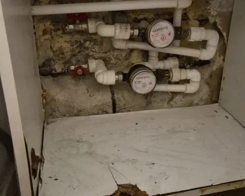O lucrare de reparație la instalația sanitară aflată într-o mască de chiuvetă cu baza deteriorată de scurgerile de apă. Se observă trasee noi din țevi PPR alb, robineți cu manetă roșie, două apometre și peretele decopertat izolat cu spumă. Pe gresie se află un clește de instalator.